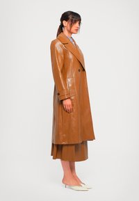 Donna in piedi di lato che indossa un cappotto in pelle marrone fino al ginocchio, una gonna abbinata, una blusa a righe e delle scarpe bianche a punta con tacco basso.