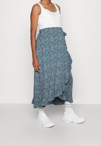 Květinová zavinovací sukně v modré, zelené a bílé barvě s volánky. Spárována s bílým tílkem a bílými sportovními teniskami.