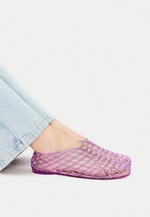 Lila Kunststoff-Slipper mit einem Gitterdesign, das offene Zehen und Fersen zeigt. Kombiniert mit hellblauen Jeans, wodurch die Transparenz des Schuhwerks hervorgehoben wird.