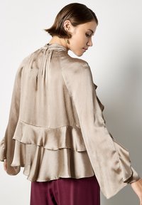 Blusa di seta beige con orlo a strati arricciato, maniche a campana lunghe e chiusura con bottone sul retro. Texture liscia con una leggera lucentezza.
