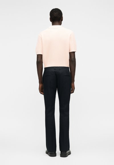 Άνδρας που στέκεται με την πλάτη, φορά ένα ανοιχτό ροζ κοντομάνικο πουκάμισο, μαύρο παντελόνι φόρμας και μαύρα παπούτσια, με φόντο μονόχρωμο.