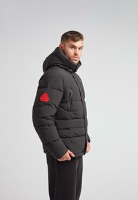 Mand iført en sort polstret jakke med hætte og rød lap på ærmet, der står sidelæns mod en simpel lys baggrund.