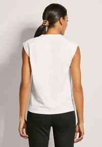 Weißes ärmelloses Oberteil mit dezenten Schulterfalten, aus weichem Stoff, kombiniert mit schwarzen, körperbetonten Hosen. Glatte Textur, schlichte Gestaltung.