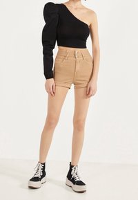 Mustapaitainen, yhdellä olkapäällä oleva toppi, jossa on puffipaita, yhdistettynä korkeavartisiin beigeihin shortseihin. Mallilla on päällä mustat korkeat tennarit, joissa on valkoiset nauhat.