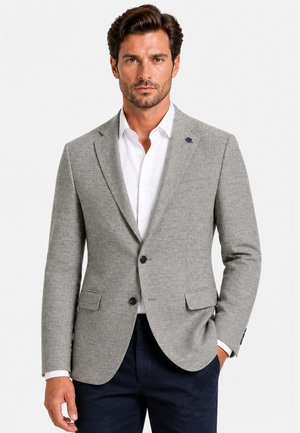 Homme portant un blazer texturé gris clair sur une chemise blanche et un pantalon foncé, debout avec une main dans la poche sur un fond blanc.
