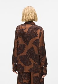 Rjava, s dolgimi rokavi srajca z listnatim vzorcem v temnejših odtenkih. Gladka tekstura in gumbi na manšetah. Rahlo ohlapen kroj.