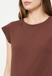 Femme portant un haut marron sans manches avec un col rond, montrant la partie supérieure du torse et une partie de son visage avec des cheveux bruns raides.