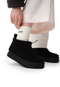 Czarne, pluszowe buty wsuwane z fakturowaną powierzchnią i grubą czarną gumową podeszwą. Widać białe skarpetki z prążkami i nadrukowanym tekstem.