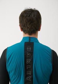 Męska kurtka sportowa w kolorze teal i czarnym, z wysokim kołnierzem, zamkiem błyskawicznym i logotypem na plecach w pionowym wzorze.