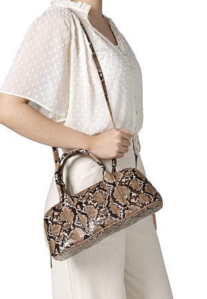 Woman wearing white textured blouse and pants holding a brown and black snake-print handbag with handles and a shoulder strap.