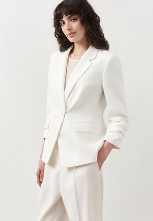 Νεαρή γυναίκα με σγουρά σκούρα μαλλιά που φοράει λευκό, ραμμένο σακάκι και ταιριαστό λευκό παντελόνι, με τα χέρια στις τσέπες, μπροστά σε απλό φόντο.