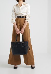 Sac cabas en cuir noir avec des accents dorés, deux poignées supérieures et une finition texturée. Associé à une chemise crème et un pantalon taille haute marron.