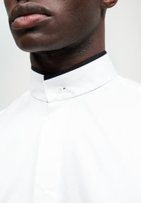 Hvit skjorte med stående krage som har en svart detalj. Stoffet ser glatt ut, med en enkel knappdetalj ved kragen.