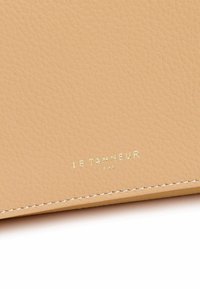 Cartera de piel color marrón claro con una superficie texturizada, que presenta un logo dorado en relieve "LE TANNEUR" y el año "1993" cerca de la parte inferior, con costuras blancas.