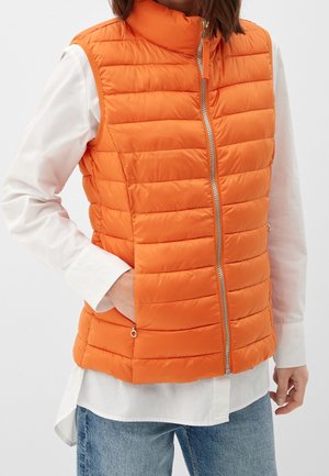 Personne portant un gilet matelassé orange vif avec fermeture éclair sur une chemise blanche à manches longues et un jean bleu, main dans la poche du gilet.