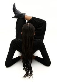 Des bottines à talons noirs sur un modèle, assorties à un haut noir à manches longues et un pantalon noir ample, avec de longues tresses qui tombent.