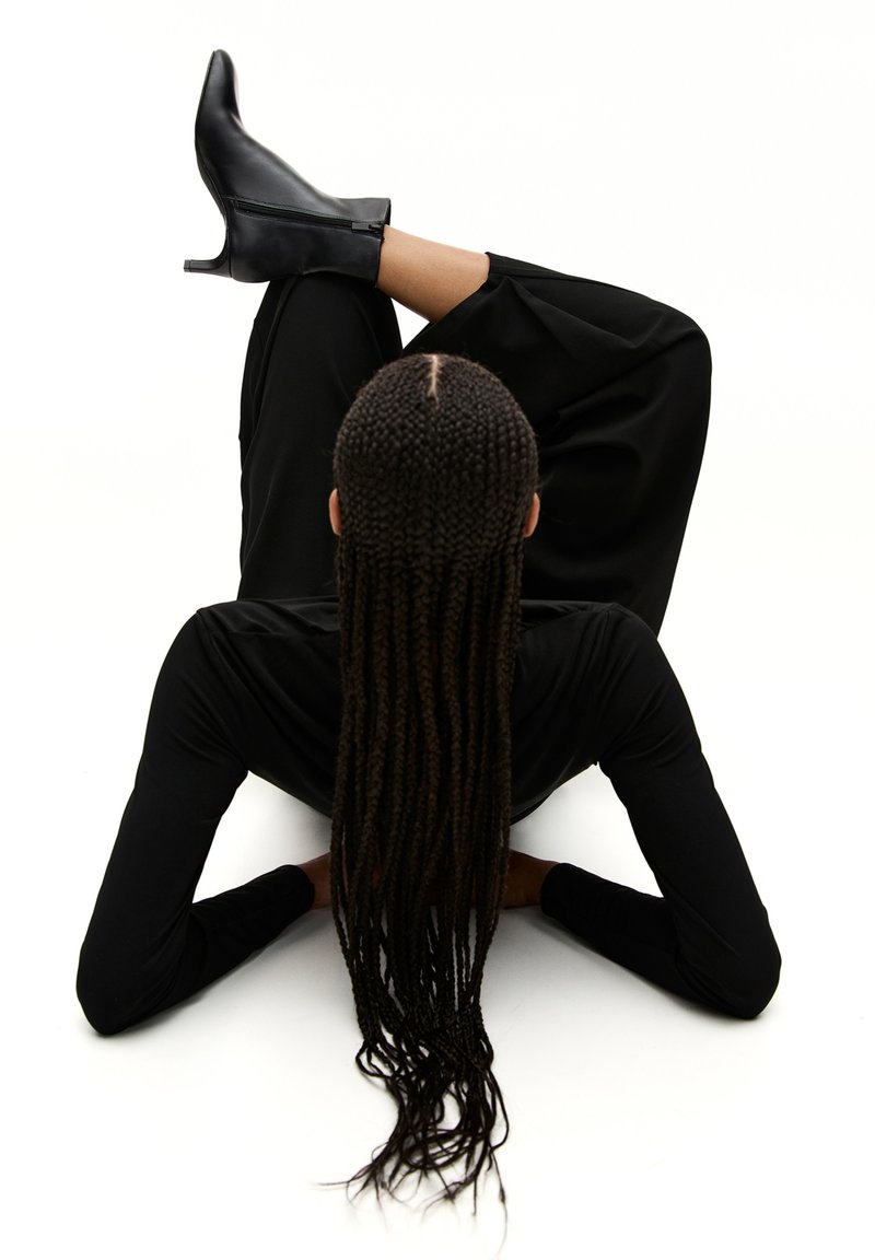Des bottines à talons noirs sur un modèle, assorties à un haut noir à manches longues et un pantalon noir ample, avec de longues tresses qui tombent.