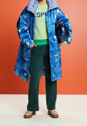 Glanzende blauwe pufferjas met een hoge kraag, gedragen over een groene trui met tekst. Donkergroene broek en beige laarzen maken de outfit compleet.