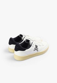 Zapatillas blancas con un acabado texturizado, con un collar negro y un emblema de calavera y huesos cruzados en gris en el costado y la lengüeta.