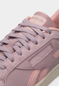 Tuva skats uz rozā Reebok sporta apavu ar auduma šņorēm un zamšādas paneļiem, izceļot zīmola logotipu sānos un mēlē.