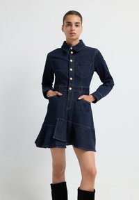 Robe en denim avec un col, fermeture à boutons sur le devant et poches plaquées. Elle présente un ourlet évasé et des coutures contrastantes en bleu foncé.