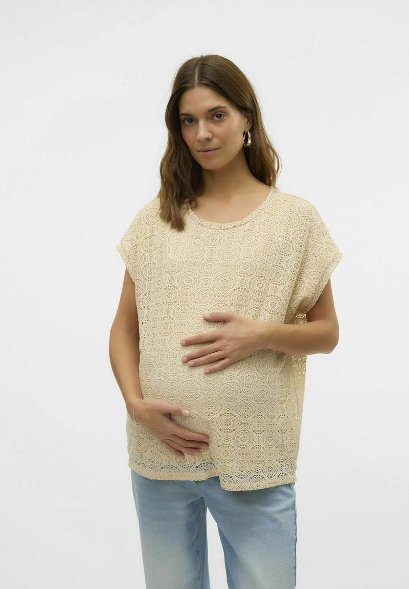 Top de encaje beige con mangas cortas, que presenta intrincados patrones circulares. Combinado con pantalones de mezclilla azul claro. La modelo sostiene una barriga visible.
