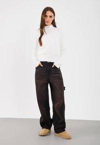 Witte geribbelde trui met ritssluiting, zwarte baggy jeans en beige instaplaarzen. Gladde stoftextuur met een aansluitend ontwerp en brede pijpen.