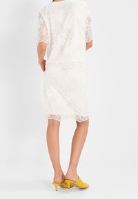 Weißes Spitzenkleid mit kurzen Ärmeln, das ein fließendes Design und subtile florale Muster aufweist. Kombiniert mit gelben High-Heels.