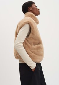 Beige Fake-Fur-Weste mit hohem Kragen, lockerer Passform und Seitentaschen, getragen über einem hellbeigen Langarmoberteil. Schwarze Hosen runden das Outfit ab.