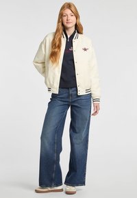 Witte bomberjack met zwart-wit gestreepte ribkraag, gedragen over een marineblauwe sweatshirt, gecombineerd met wijde blauwe jeans en sneakers.