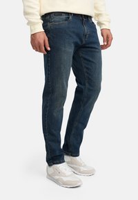 Man draagt donkerblauwe spijkerbroek, crèmekleurige trui en witte sneakers, staand tegen een effen lichte achtergrond.