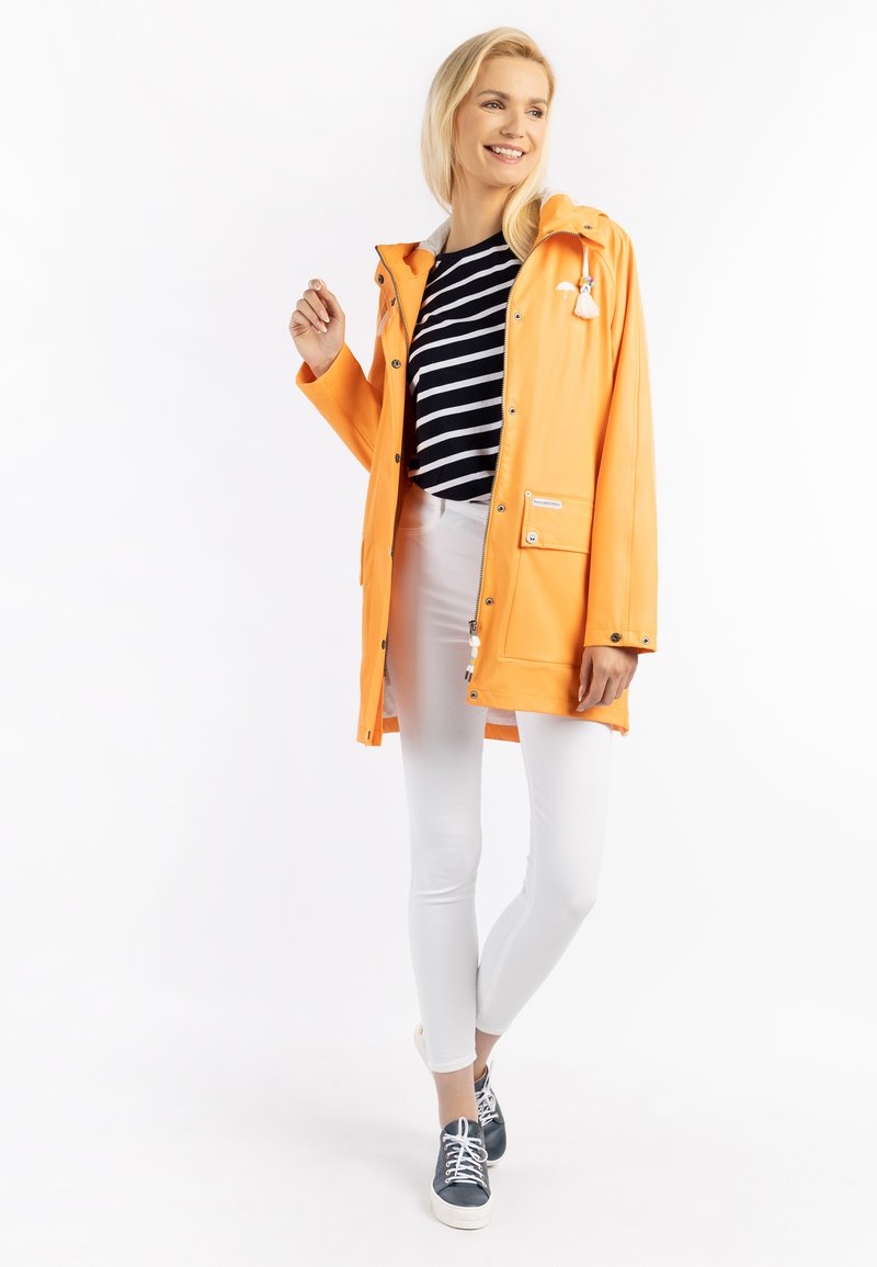 Narancssárga vízálló esőkabát kapucnival, cipzárral záródik, oldalzsebekkel rendelkezik, alatta pedig egy csíkos fekete-fehér póló van.