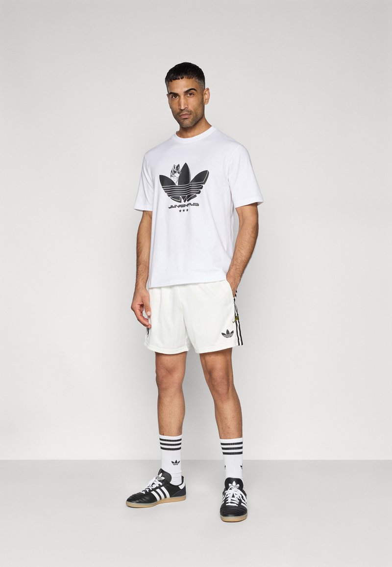 Weißes T-Shirt mit schwarzem Grafiklogo, weiße Shorts mit schwarzen Seitenstreifen, schwarze Sportschuhe und gestreifte Socken. Einfaches Design, legere Passform.