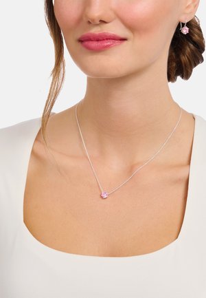Silberne Kette mit einem Anhänger aus pinkfarbenem Edelstein; passende pinkfarbene Ohrringe; getragen zu einem weißen Oberteil mit weitem Ausschnitt.
