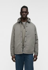 Γκρι επενδυμένο μπουφάν με όρθιο γιακά, κουμπώματα με πατήστε και εμπρόσθιο φερμουάρ. Φορώντας πάνω από γκρι πουκάμισο, συνοδευόμενο από μπλε παντελόνι.