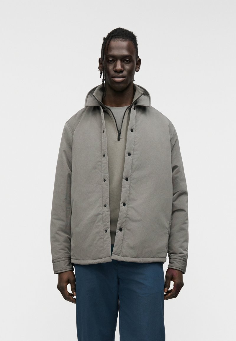 Γκρι επενδυμένο μπουφάν με όρθιο γιακά, κουμπώματα με πατήστε και εμπρόσθιο φερμουάρ. Φορώντας πάνω από γκρι πουκάμισο, συνοδευόμενο από μπλε παντελόνι.