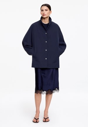 Kvinde med mørkt hår sat op i en lav knold iført en navy oversized jakke over en navy midi-nederdel med blonder og sorte sandaler med stropper, stående.