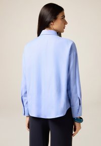 Camisa de manga larga azul claro hecha de tela suave, con cuello clásico, aberturas en los puños y dobladillo ligeramente redondeado.