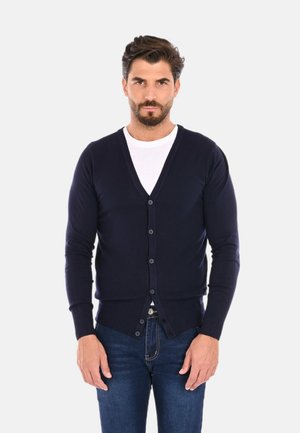 Cardigan lavorato a maglia blu navy con scollo a V, chiusura a bottoni e polsini a coste. Indossato sopra una maglietta bianca, abbinato a jeans blu scuro.