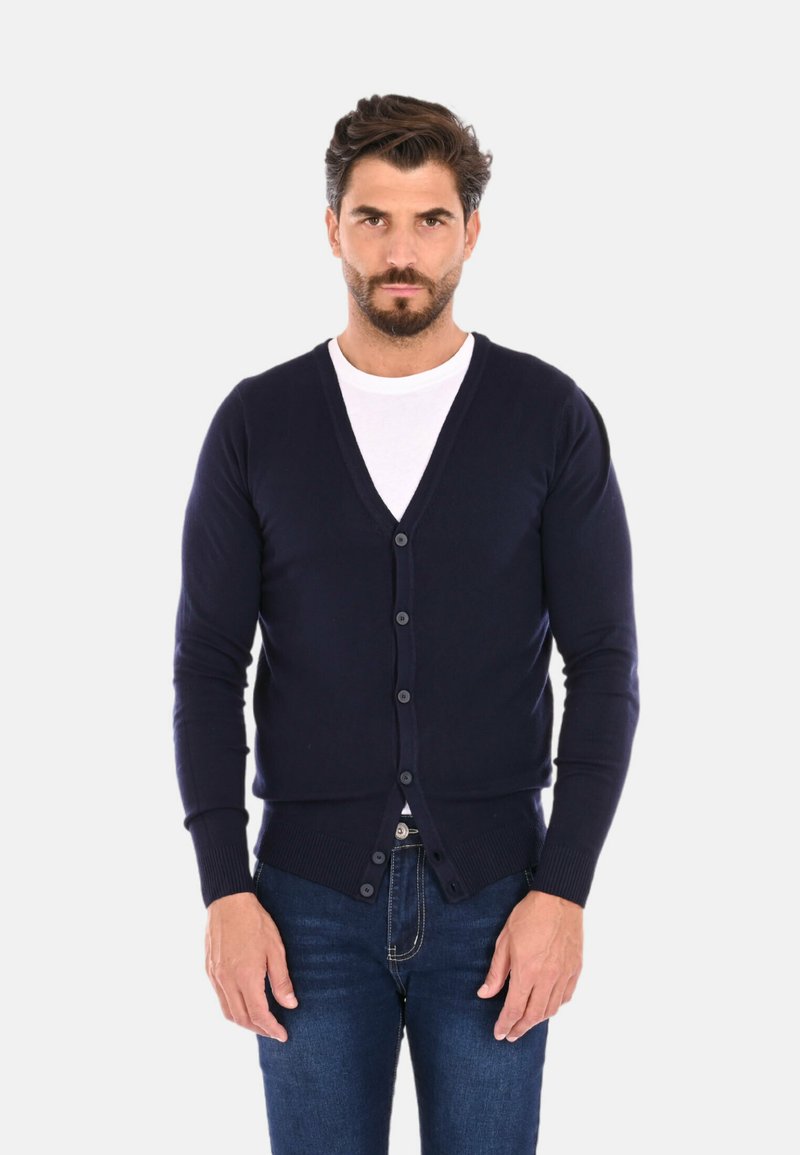 Cardigan lavorato a maglia blu navy con scollo a V, chiusura a bottoni e polsini a coste. Indossato sopra una maglietta bianca, abbinato a jeans blu scuro.