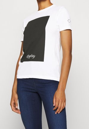 Persona vistiendo una camiseta blanca de manga corta con un gran cuadrado negro y el texto "Replay", combinada con jeans azul oscuro, sobre un fondo sencillo.