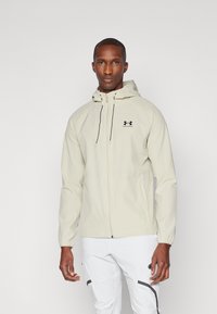Giacca a zip beige chiaro con cappuccio, dotata di polsini elasticizzati e di un logo sul petto, realizzata in materiale liscio e resistente alle intemperie.