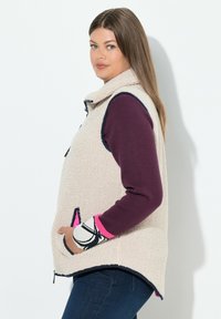 Fleeceweste in Creme mit hohem Kragen, marineblauen Akzenten und Taschen. Die Ärmel in tiefem Lila weisen pinke und schwarze grafische Bündchen auf.