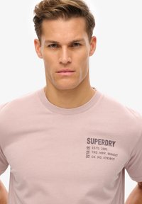 Lyserød bomulds t-shirt med rund halsudskæring, korte ærmer og grå trykt branding på forsiden med tekstdetaljer.