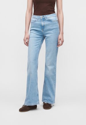 Personne portant un jean évasé taille haute bleu clair et des chaussures marron, debout devant un fond clair uni.