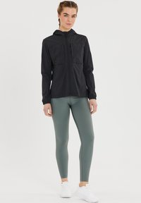 Kvinde står i atletisk outfit med sort hættejakke, grå tights og hvide sneakers på en ensfarvet hvid baggrund.