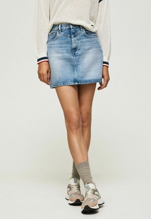 Vrouw die een lichtblauwe denim mini-rok draagt, met beige sokken en beige-grijze sneakers, staand met gekruiste benen.