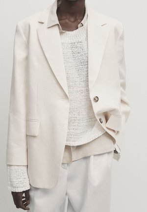 Personne porte un blazer crème oversize sur un pull tricoté blanc et une chemise beige clair, associé à un pantalon crème ample sur un fond uni.
