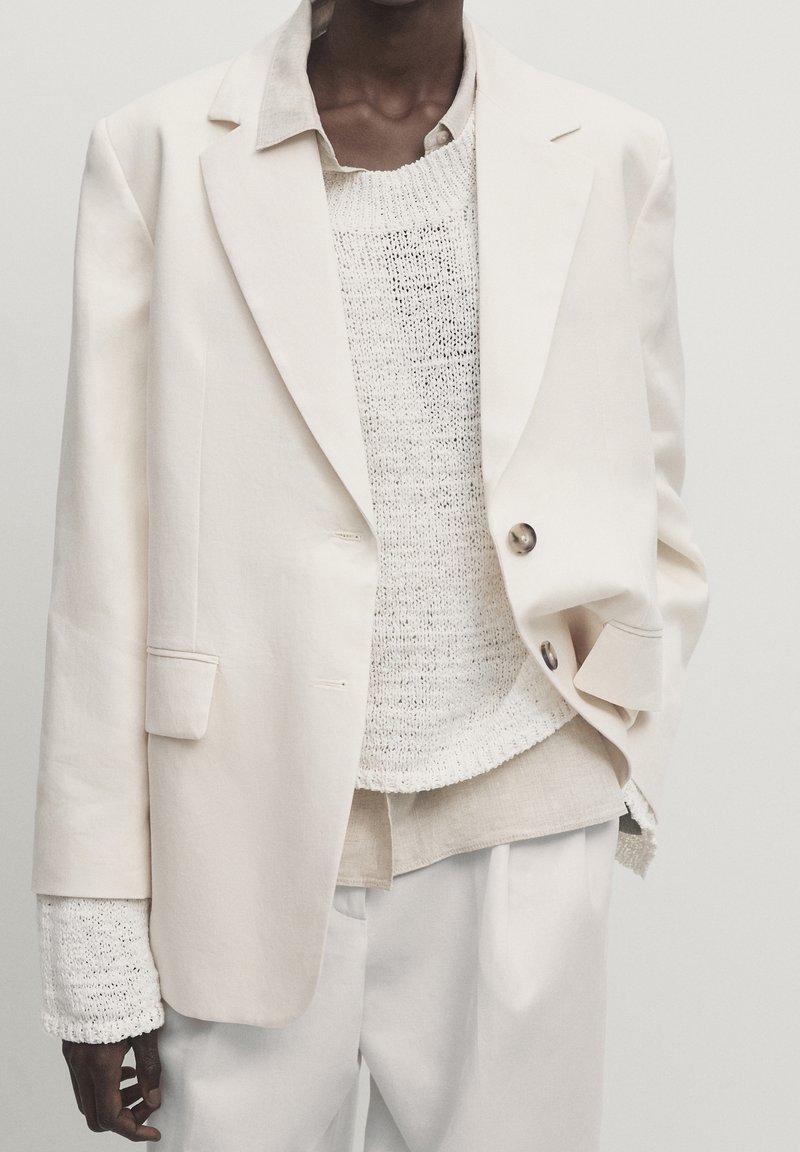 Personne porte un blazer crème oversize sur un pull tricoté blanc et une chemise beige clair, associé à un pantalon crème ample sur un fond uni.