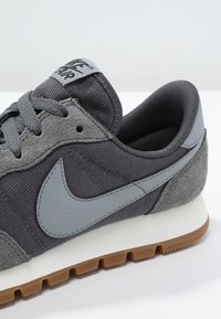 Le sneaker Nike grigie presentano un mix di tessuto testurizzato e suede. Il design comprende un logo swoosh grigio e una suola in gomma con accenti marroni.